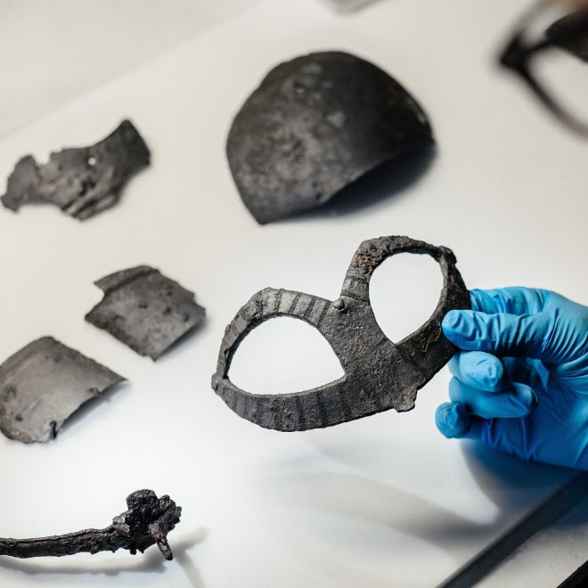 Nærbilde av en hånd som holder i en del av en vikinghjelm, flere vikinggjenstander ligger på et bord.