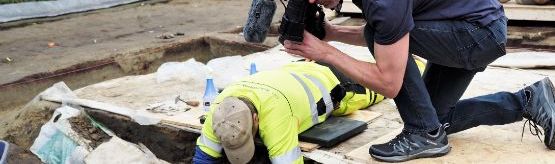 Close up of a man taking a picture of a man working at an excavation area.